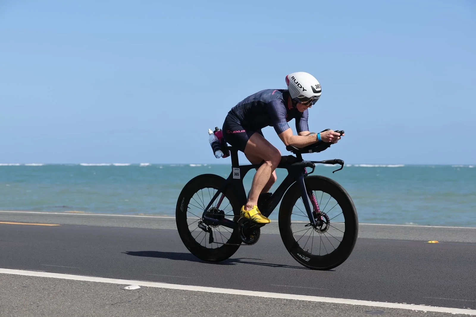 A cyclist riding a black bike along a coastal road over water, wearing a white helmet, dark cycling attire, and yellow shoes.